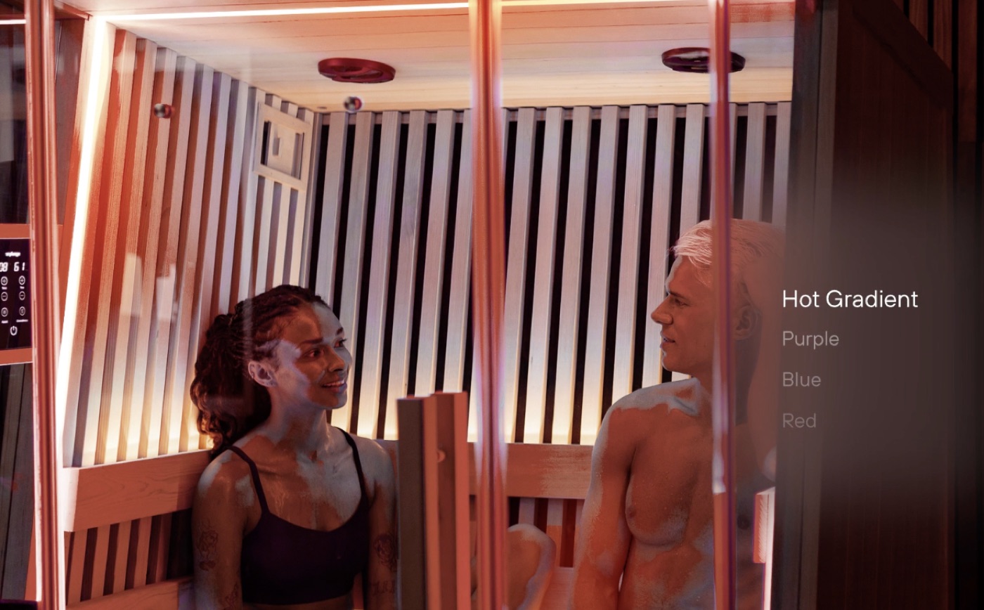 Two people talking inside a slatted-wood infrared sauna lit in warm orange, with a lighting colour menu at the right